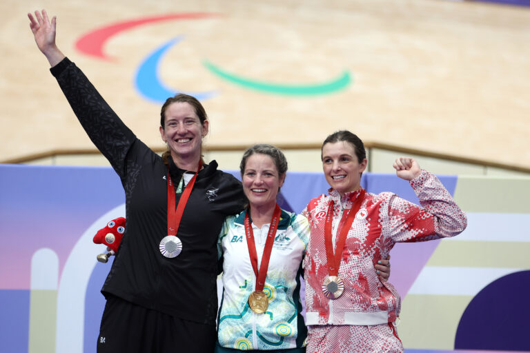 Anna Taylor celebrates her silver medal on the podium following the Women’s C4 3000m Individual Pursuit at the Paris 2024 Paralympic Games. The Paralympic Agitos logo can be seen on the track behind and Anna raises her hand in celebration.