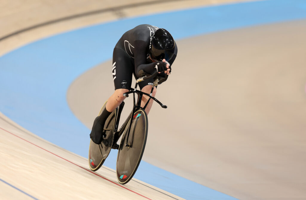 Devon Briggs cycles on the velodrome during the Men's C1-3 1000m Time Trial Qualifying. He wears a black NZ Paralympic Team uniform.