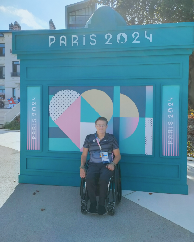 Paralympian #126 Tim Johnson poses at the Paris 2024 Paralympic Games in front of a promotional banner. He attended as a Wheelchair rugby official. 