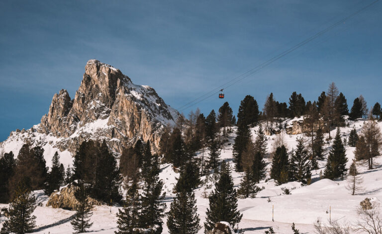 A snowy skiing mountain with a jagged rocky peak is covered in dark pine trees. A gondola ascends the mountain to the right.