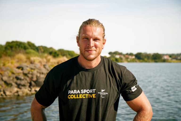 Portrait of Para athlete Finn Murphy on the shore of a lake. He has long blonde hair worn tied back. He wears a black t-shirt with white and yellow text reading "Para Sport Collective."