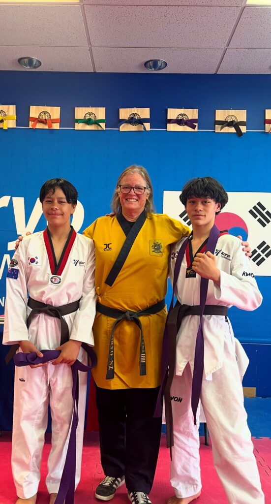 Para taekwondo coach Camille Pruckmuller stands in a gold uniform with a black belt. Beside her stand two young taekwondo athletes in white uniforms and purple belts.