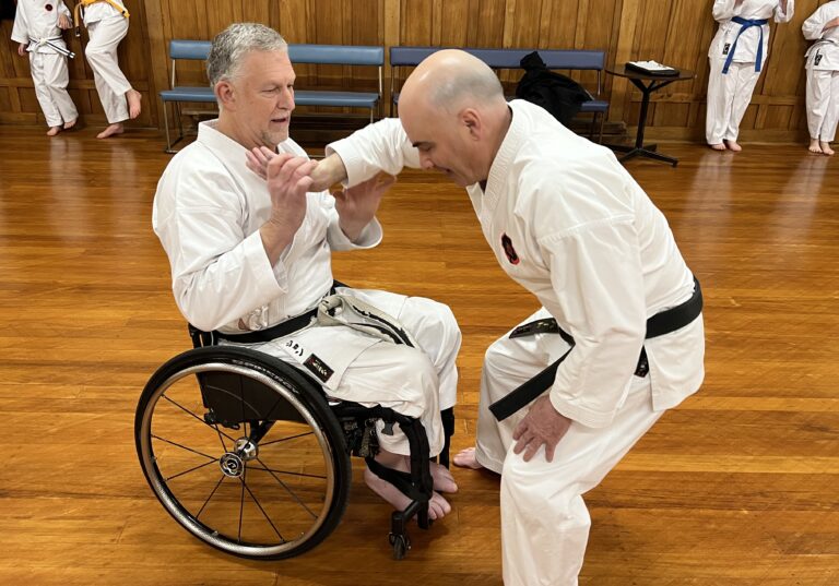 John Marrable teaches karate in his wheelchair. He wears a white uniform with a black belt.