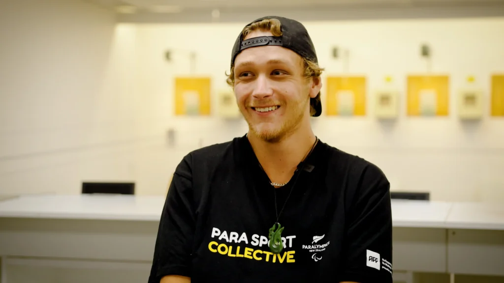 A photo of Eligh Fountain smiling in front of a target shooting range. He is wearing a Para Sport Collective T-shirt.