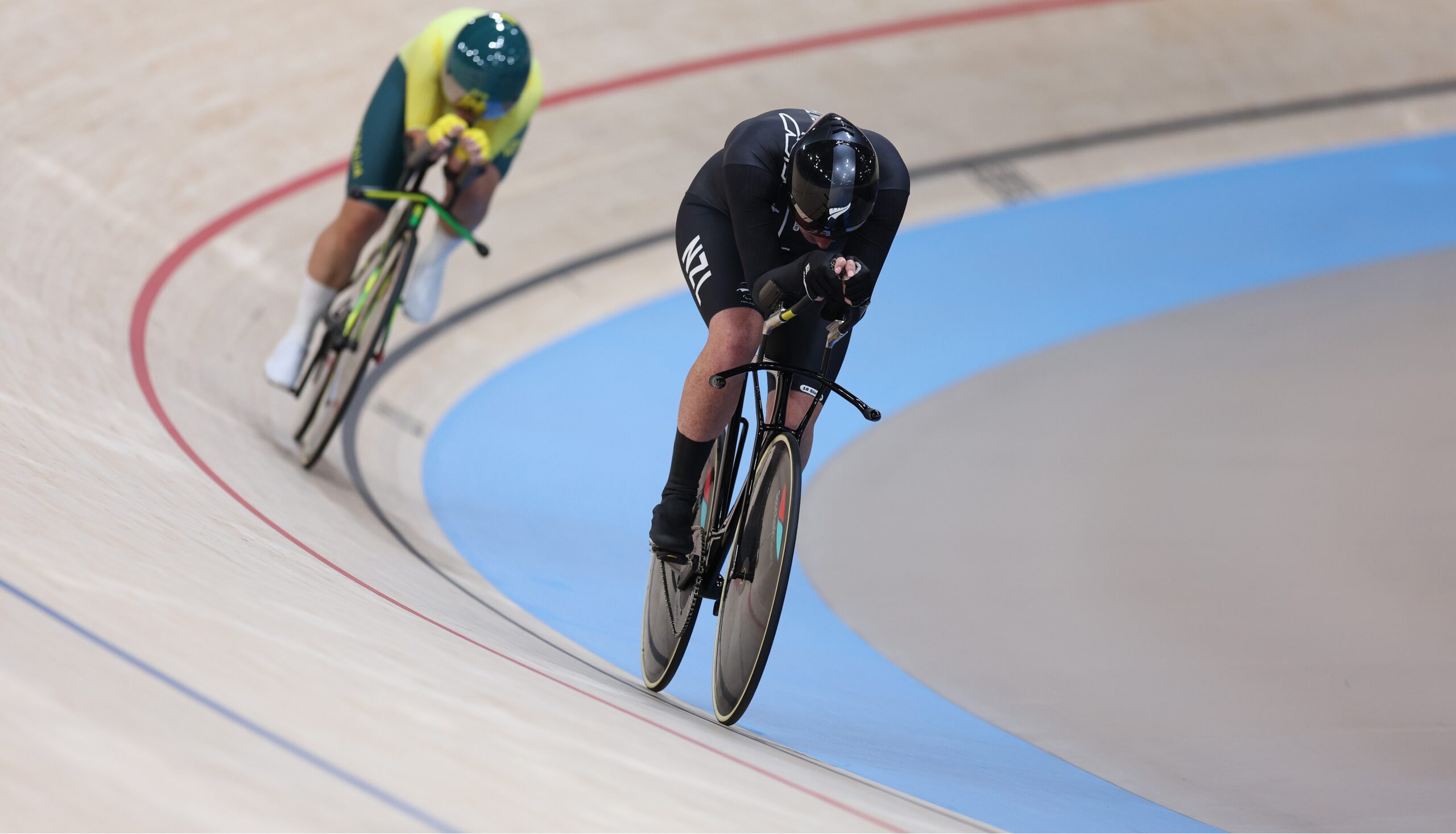 Paralympian #225 Anna Taylor cycles on a velodrome at the Paris 2024 Paralympic Games. An Australian Para cyclist is behind her.