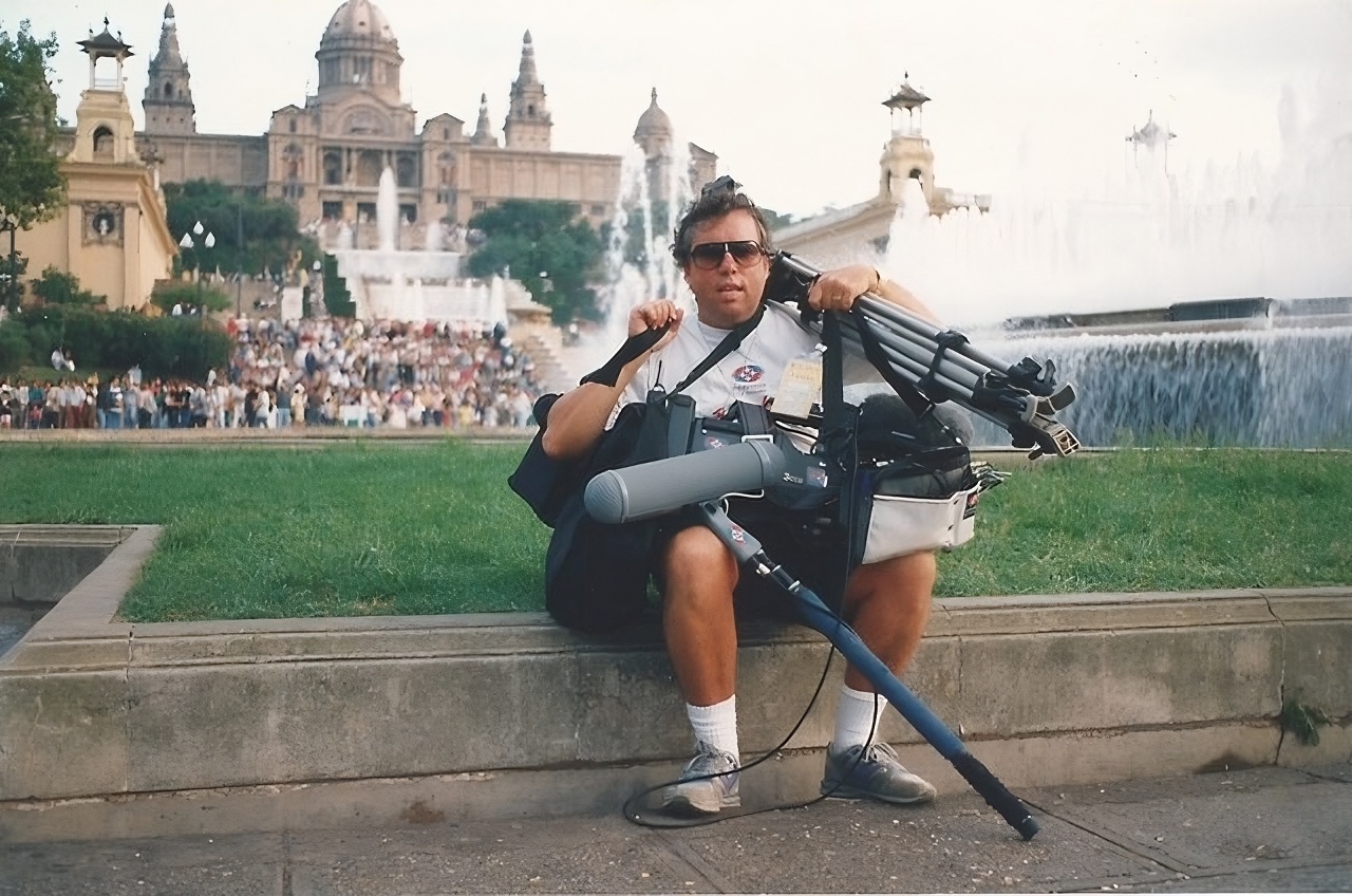 Chas Toogood sits on a ledge in a Barcelona park carrying a range of filming equipment, including a boom microphone, a tripod and a bag.
