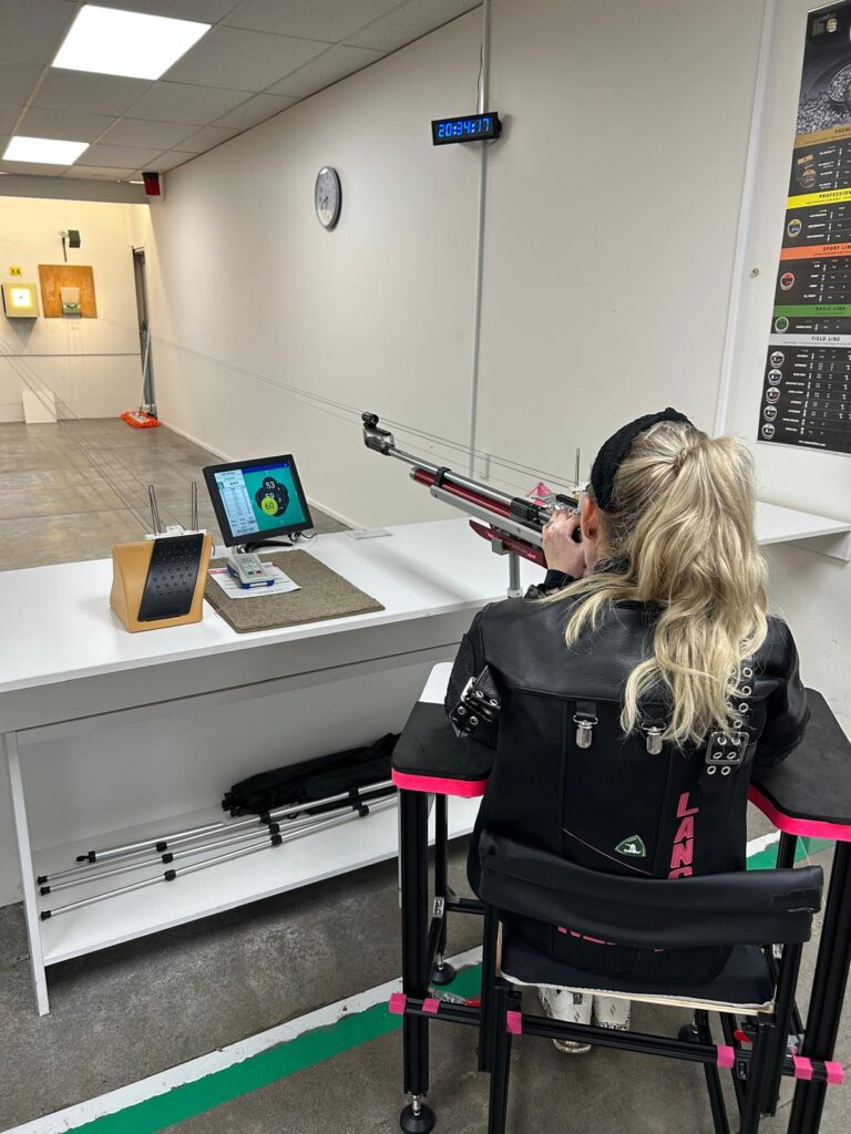 Liz aims her rifle. Her shooting jacket has her name "LANGDON" stitched onto the back in bright pink letters.