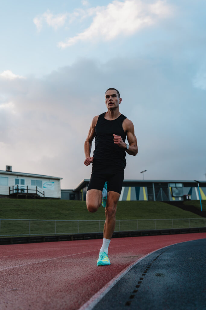 A low wide angle photo of Michael Whittaker running on an athletics track under a cool evening light.