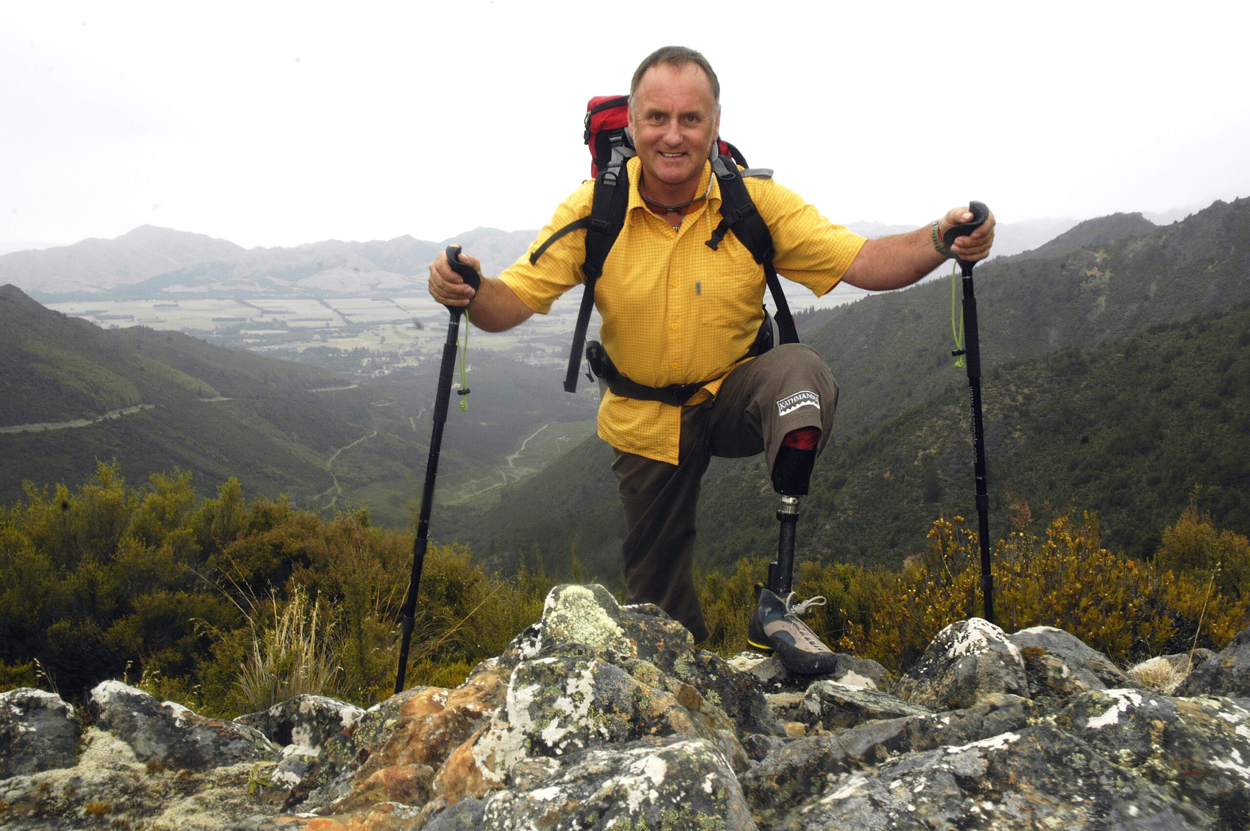 A photo of Mark Inglis at the top of a mountain. He holds two hiking poles and wears a prosthetic leg.