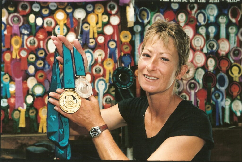 Jayne Craike holds her two medals in front of a wall of ribbons.