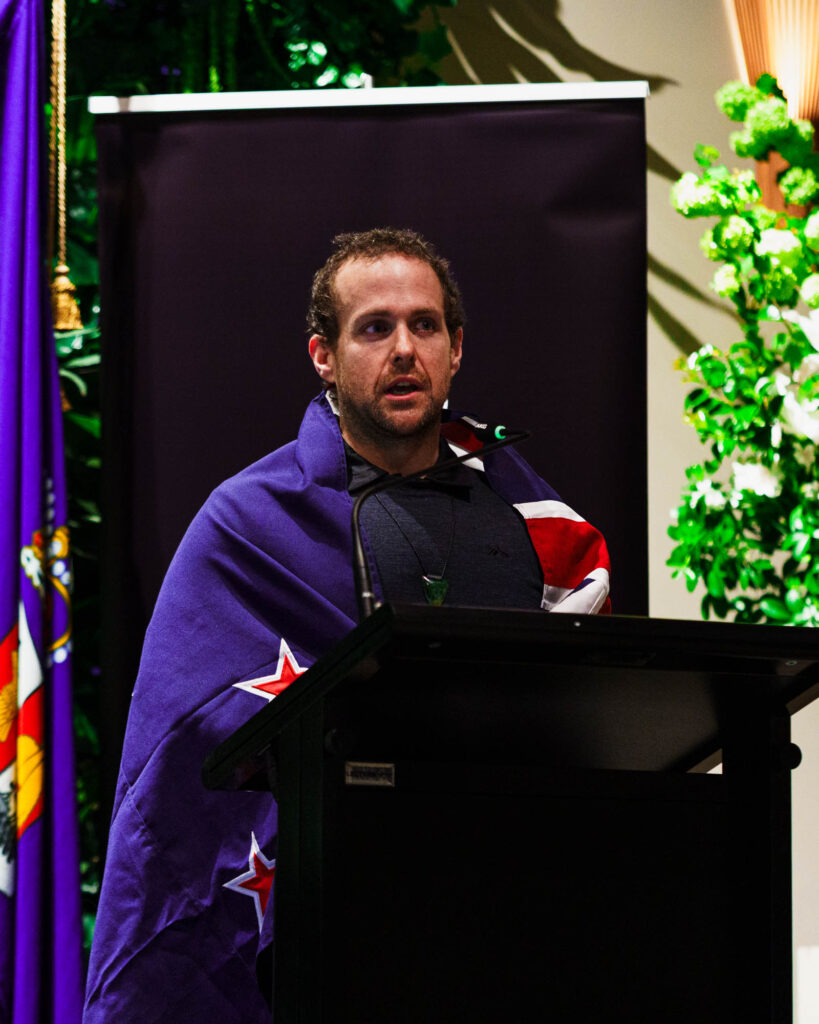 Paralympian #158 Adam Hall speaks behind a lectern with a NZ flag wrapped around himself.