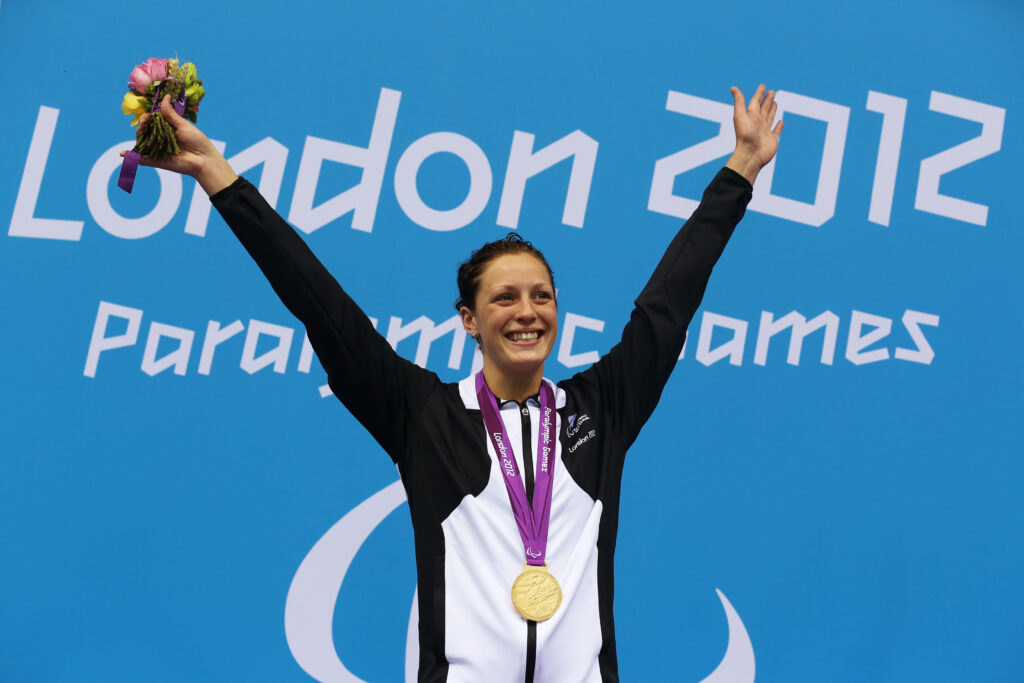 Sophie Pascoe raises her hands in celebration with a gold medal around her neck. A blue background shows the London 2012 Paralympic Games logo.