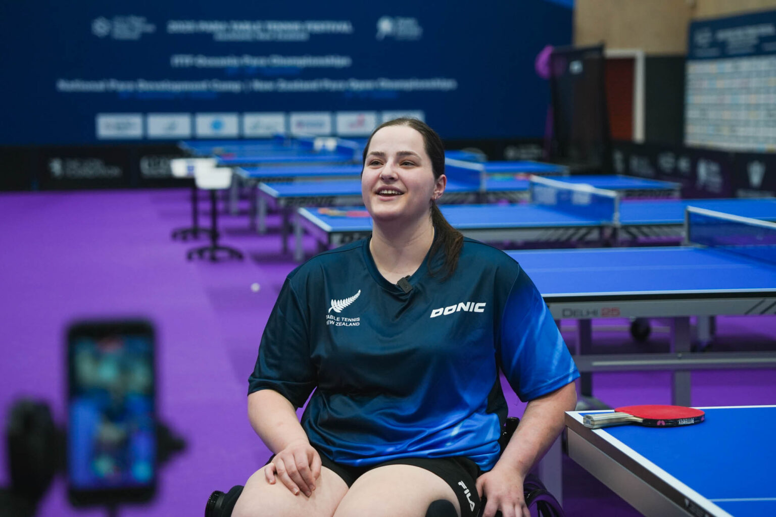 Sarah Clarke laughs during an interview in front of a line of table tennis tables.