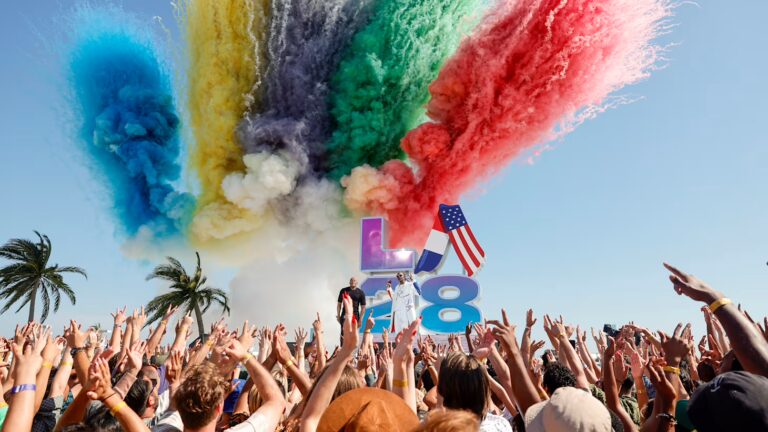 A crowd cheers in front of a stage with the LA28 logo. Colourful flares launch in the background.