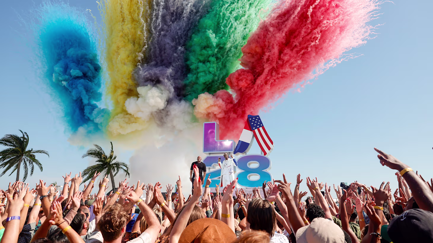 A crowd cheers in front of a stage with the LA28 logo. Colourful flares launch in the background.