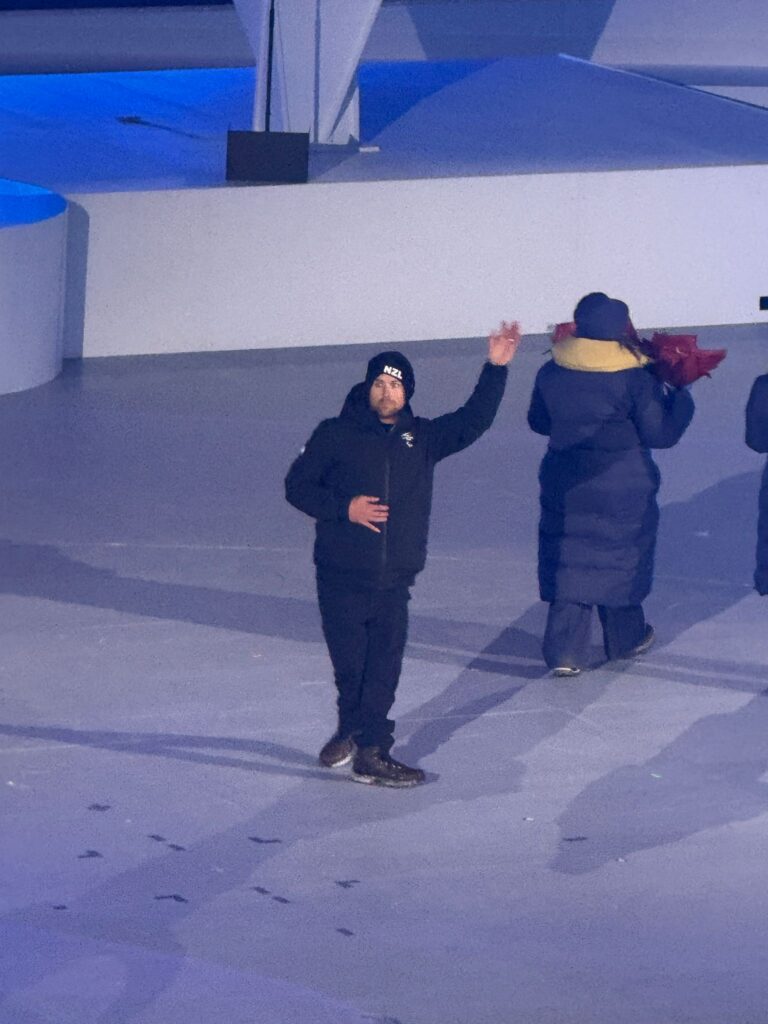 A man with his hand up in a wave turned temporarily from following a line of people carrying flowers