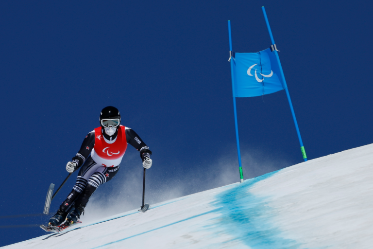 Adam Hall competes on the slopes in a black Kiwi gear with a red bib. On the right-hand side is a blue coloured gate.