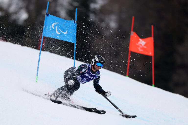 Paralympian #158 Adam Hall MNZM on the slopes at the Milano Cortina 2026 Paralympic Winter Games in the Men's Giant Slalom Standing. He is carving a turn on his skis in black coloured clothing with blue and red coloured gates behind him.