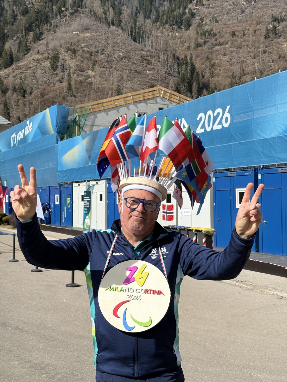 Volunteer at Paralympic Village with a hat holding multiple country flags and a big Milano Cortina 2026 sign around his neck