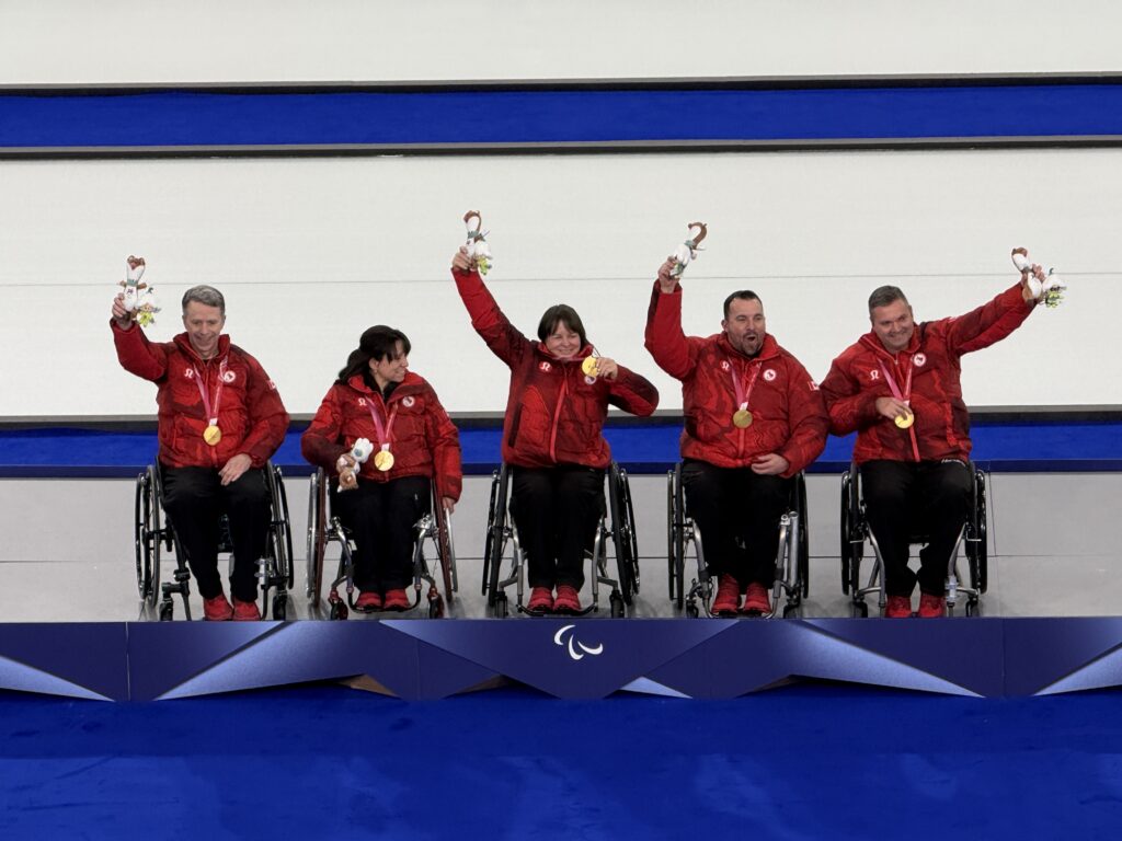 team of wheelchair curling athletes wearing medals and holding up mascots