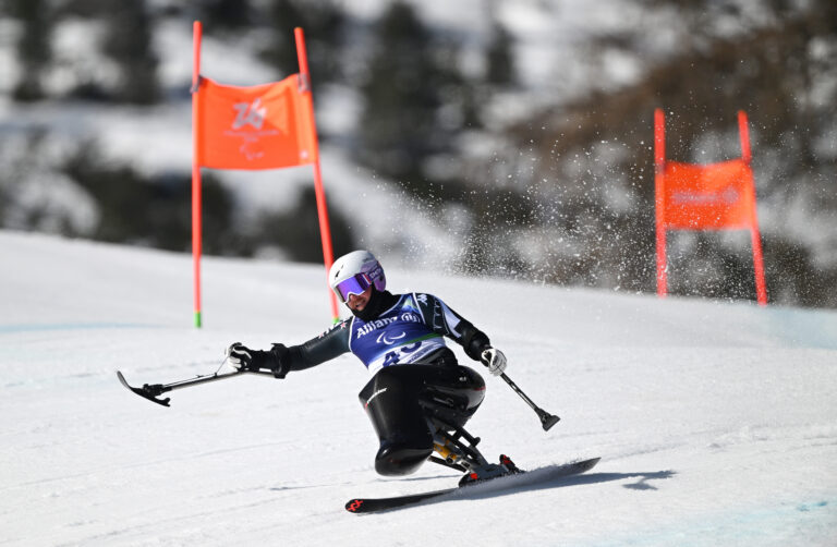 Sit skier Corey Peters in mid-race action in the Men's Downhill Sitting at the Milano Cortina 2026 Paralympic Winter Games. He has wearing in NZ Paralympic Team colours and is competing at high speed between the gates.