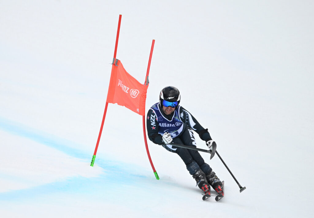 Para alpine skier Adam Hall in action in the Men's Giant Slalom Standing. He is dressed in black kit on his skis passing a red gate to the left hand side.