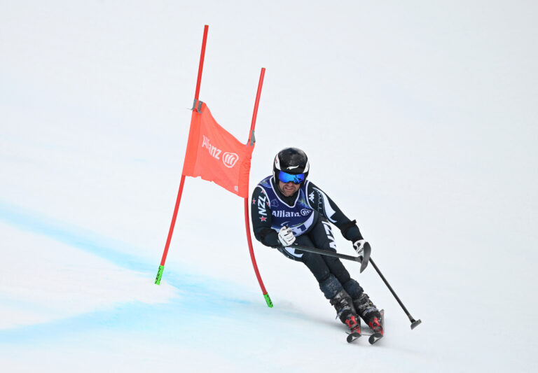 Para alpine skier Adam Hall in action in the Men's Giant Slalom Standing. He is dressed in black kit on his skis passing a red gate to the left hand side.