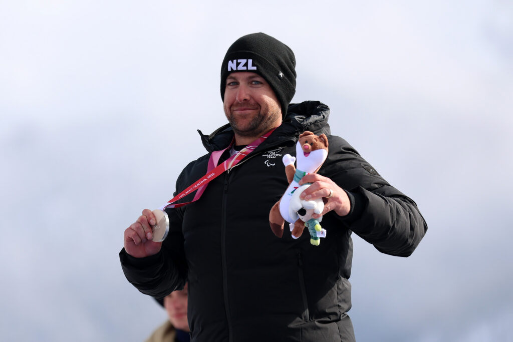 Adam Hall in a New Zealand beanie and jacket on the medal podium with a silver medal in his right hand and a mascot in the left hand.