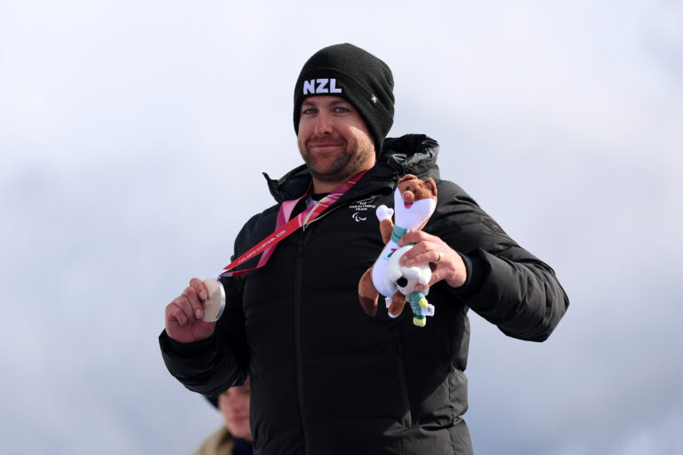 Adam Hall in a New Zealand beanie and jacket on the medal podium with a silver medal in his right hand and a mascot in the left hand.