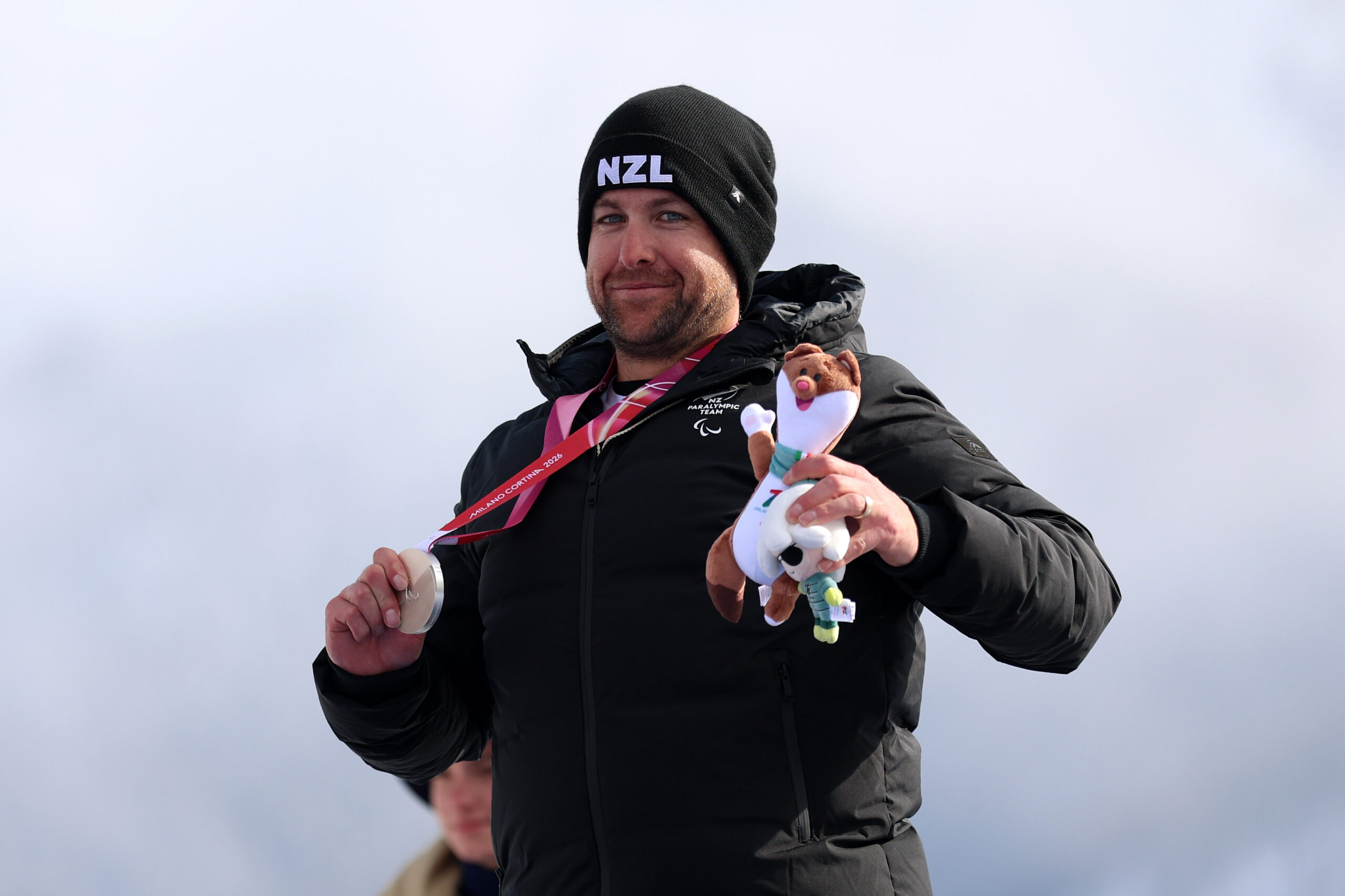 Adam Hall in a New Zealand beanie and jacket on the medal podium with a silver medal in his right hand and a mascot in the left hand.