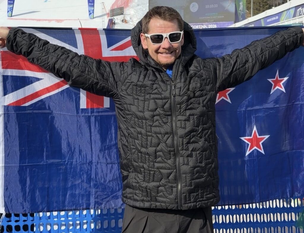 Paralympian #232 Wojtek Czyz in a black jacket and sunglasses with arms outstretched carrying the New Zealand flag at the Milano Cortina 2026 Paralympic Winter Games.