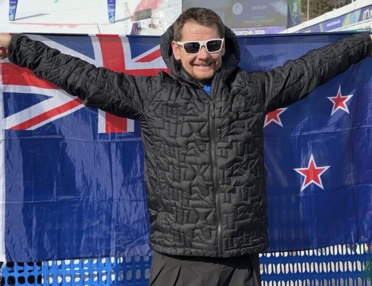 Paralympian #232 Wojtek Czyz in a black jacket and sunglasses with arms outstretched carrying the New Zealand flag at the Milano Cortina 2026 Paralympic Winter Games.