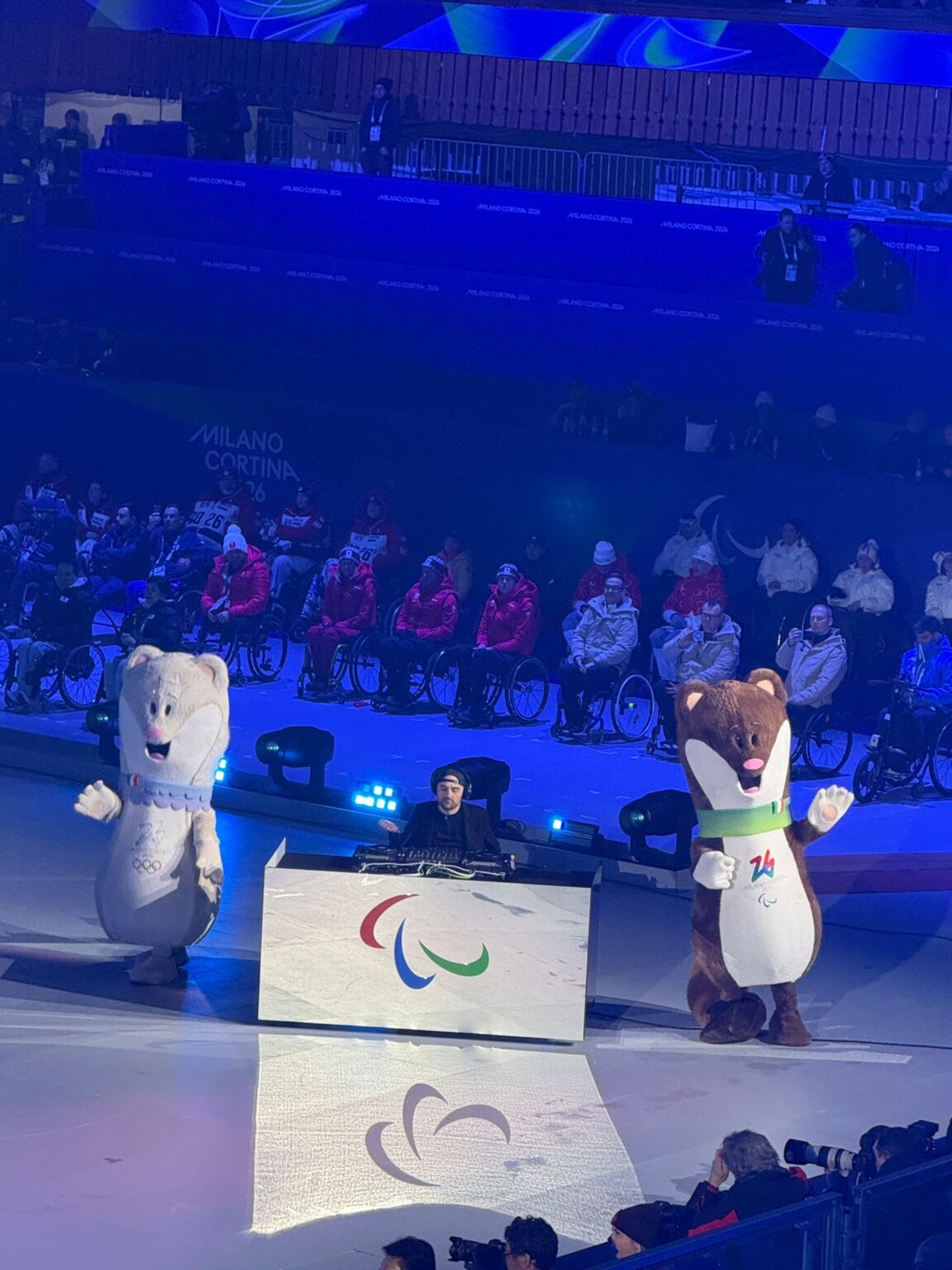 A DJ operating at a table with the Paralympic agitos, and the two mascots. People in wheelchairs in the background.