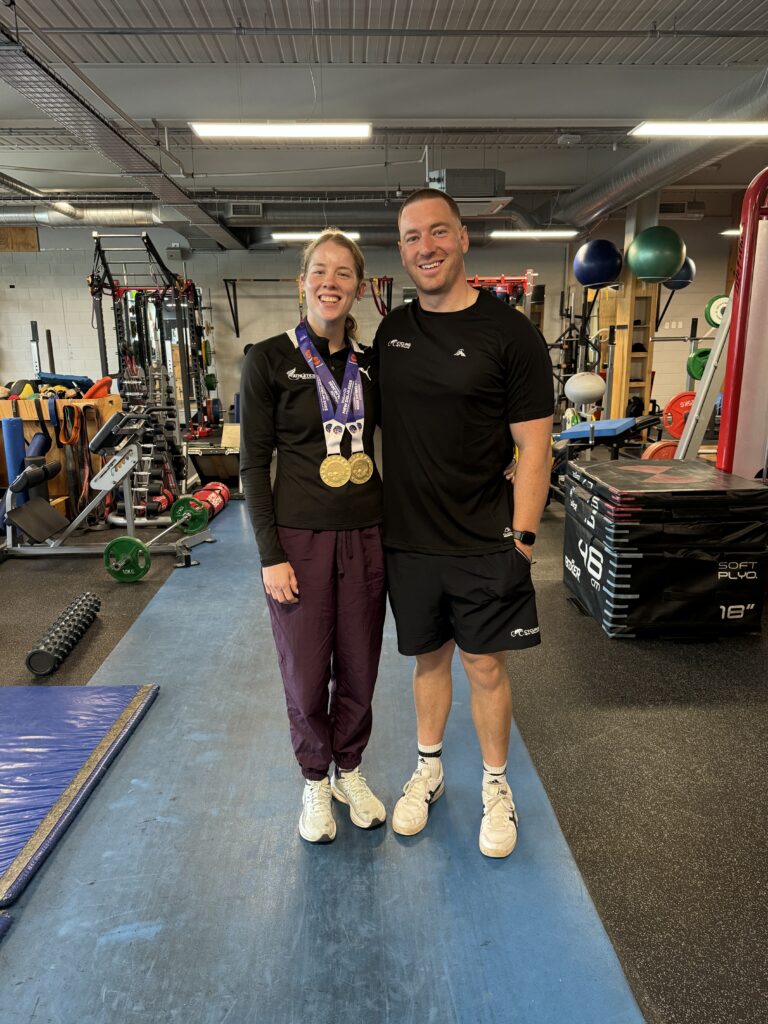 Danielle stands with coach George in a gym, wearing two gold medals.