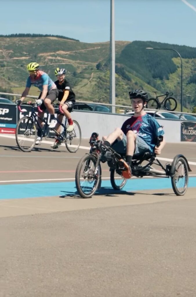 A young cyclist rides a seated bike. A tandem bike is seen in the background. 