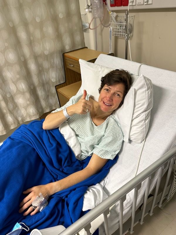 Helene lies in a hospital bed, smiling holding a thumbs up.