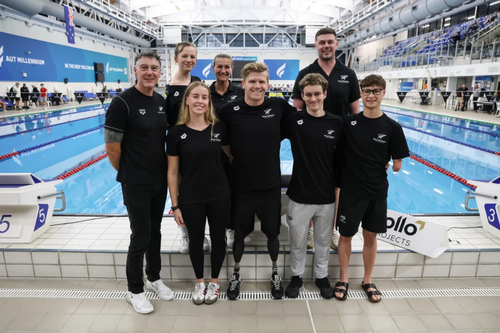 Five NZ Para swimmers stand poolside with coaches.