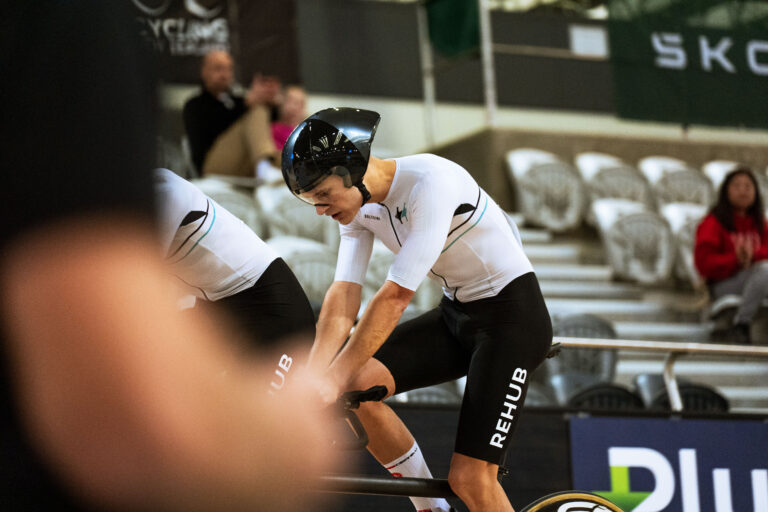Michael Whittaker sits in the stoker position on a tandem bicycle on track at a velodrome.